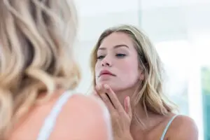 Focused beautiful young woman looking at herself in the bathroom mirror at home, concerned about acne.