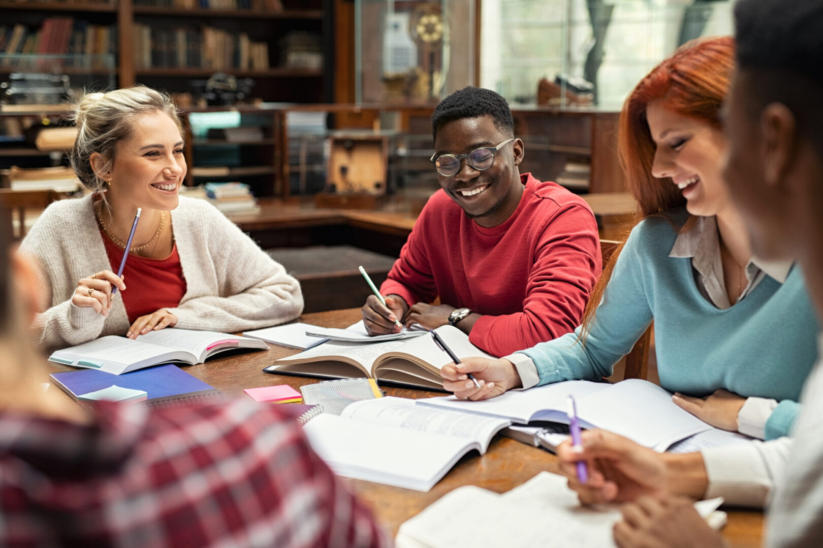 Happy college students studying together and laughing. Group of multiethnic friends smiling and studying while sitting in university library, representing great cognitive ability.