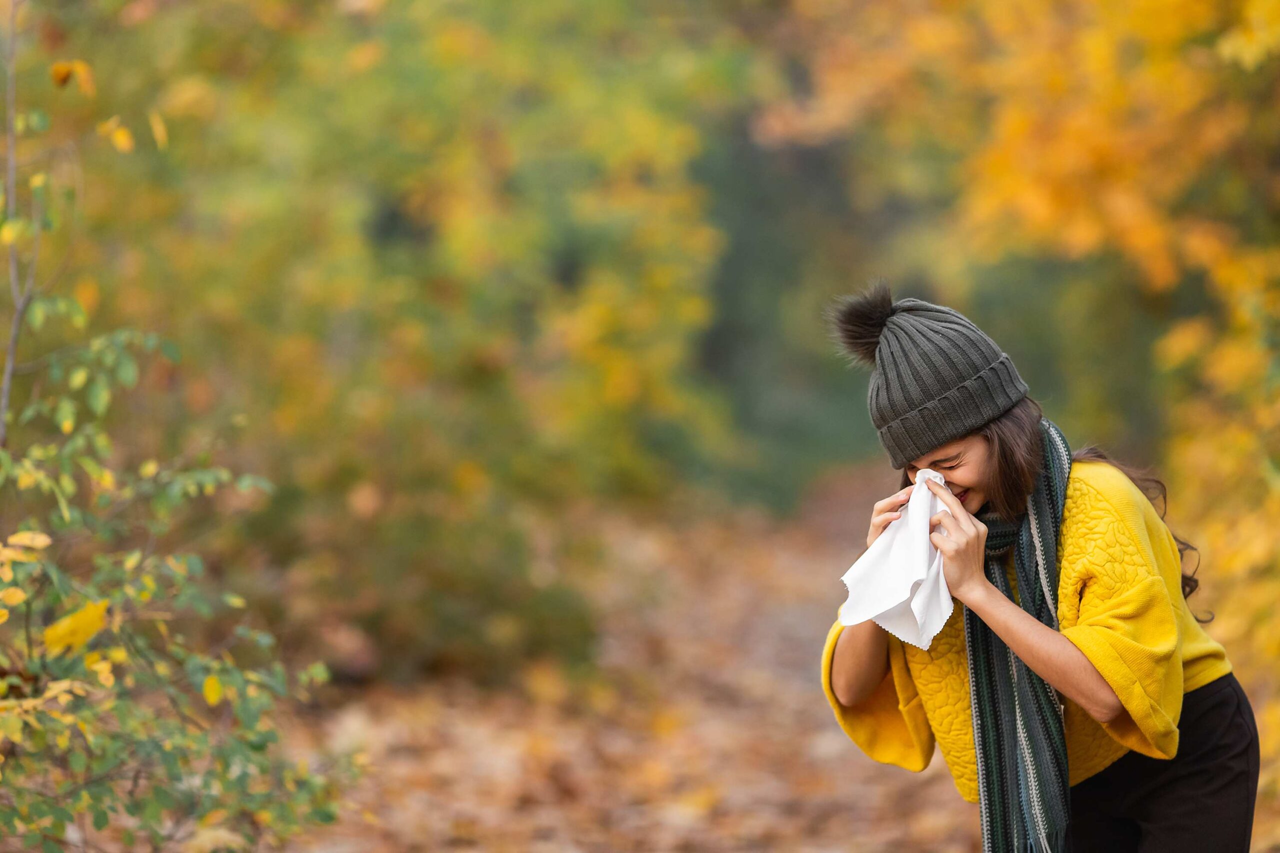 Woman sneezing outdoors with colorful autumn leaves in the background, showing why many people explore quercetin for allergies in fall.