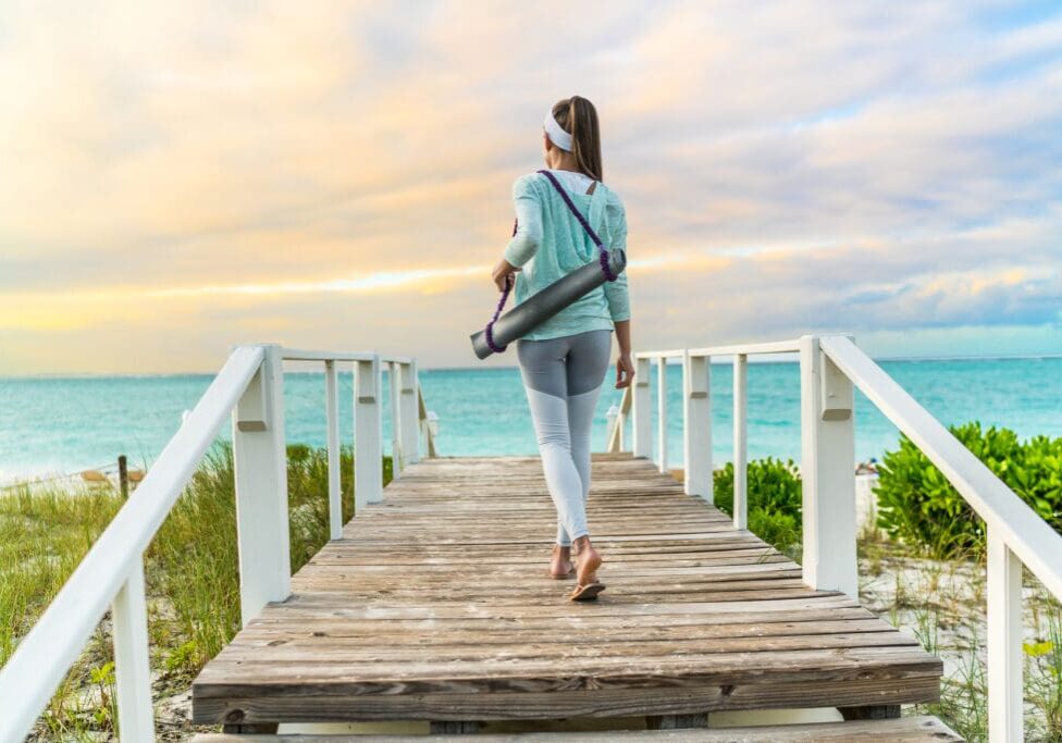 Woman walking to beach on a boardwalk carrying yoga mat for healthy methylation