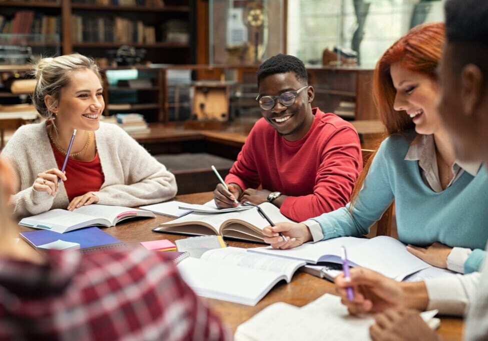 Happy college students studying together and laughing. Group of multiethnic friends smiling and studying while sitting in university library, representing great cognitive ability.