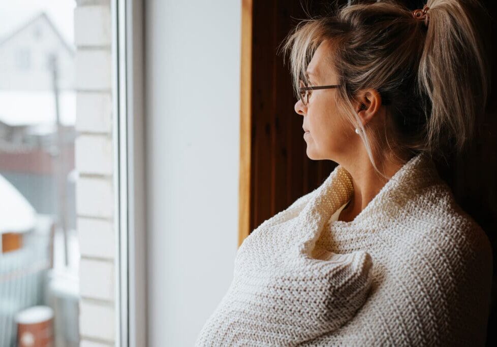 Side view portrait of sad lonely adult caucasian woman wrapped in warm cozy knitted blanket standing by window and looking away, indoors. Winter blues.