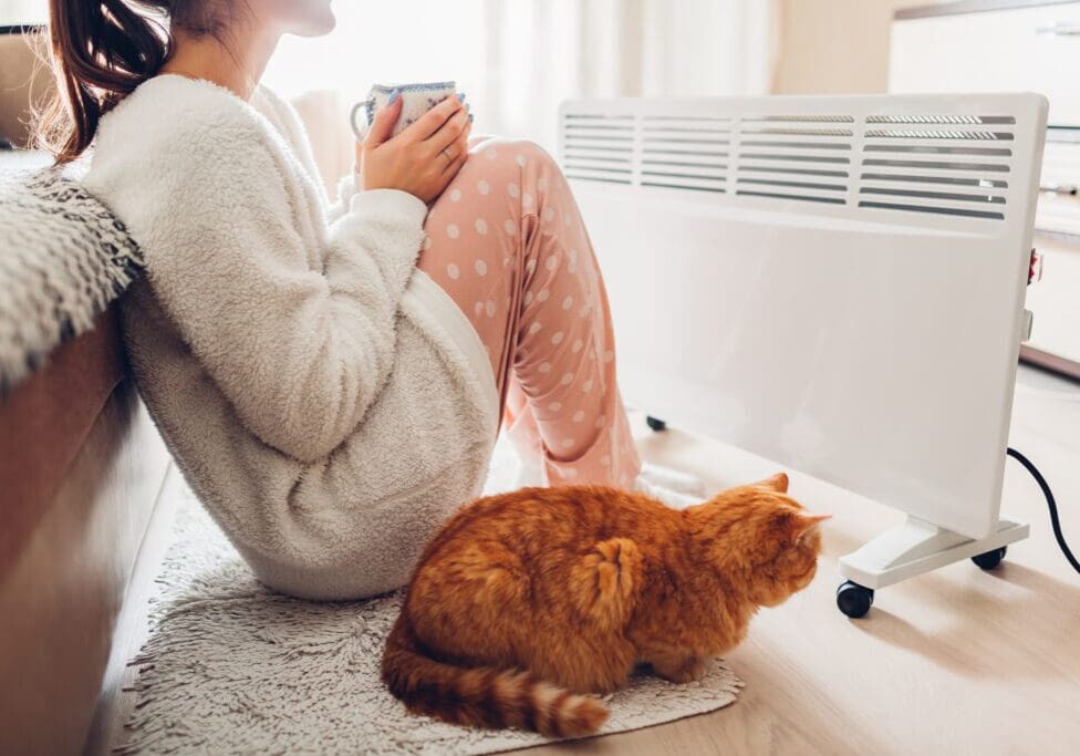 Using heater at home in winter. Woman warming and drinking tea with cat.